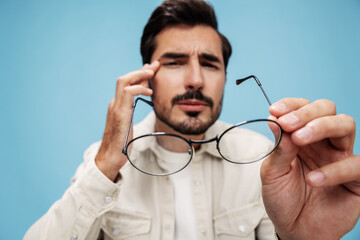 Close-up portrait of a brunette man looking through glasses that he holds in his hands, eye problems, glasses for vision farsightedness and nearsightedness, on a blue background, copy space 