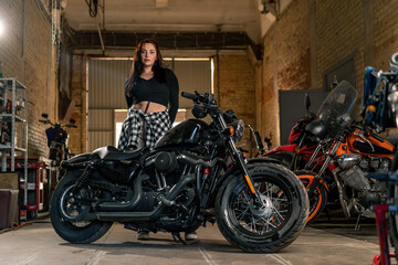 Plakat Creative authentic motorcycle workshop garage beautiful young biker girl standing near a cool motorcycle
