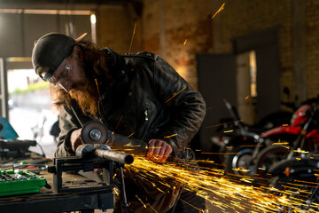 Fototapeta na wymiar Creative authentic motorcycle workshop redhead bearded brutal biker works with circular saw Sparks fly from hot metal