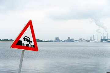 Verkehrsschild Achtung Ufer links im Bild, im Hintergrund defokussiert Wasser und Hafengebäude zu sehen, horizontal