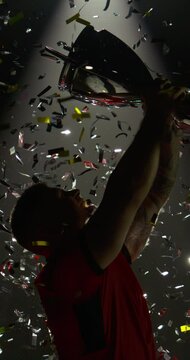 VERTICAL VIDEO - Silhouette of Caucasian male rugby player raising a trophy above head against bright light and falling confetti. Super slow motion, shot on RED cinema camera