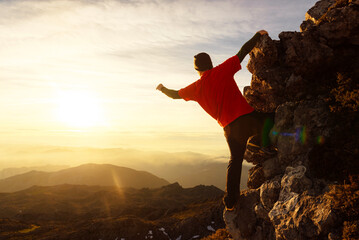 man mountaineer and hiker perched on a mountain rock contemplating the beautiful sunset and raising...