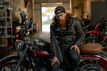 Obraz na płótnie Canvas Creative authentic motorcycle workshop Garage brutal serious bearded biker mechanic sitting on motorcycle