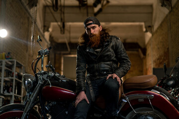 Obraz na płótnie Canvas Creative authentic motorcycle workshop Garage brutal serious bearded biker mechanic sitting on motorcycle