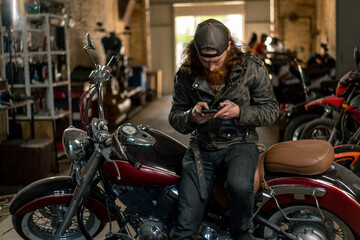 Obraz na płótnie Canvas Creative authentic motorcycle workshop Garage brutal serious bearded redhead biker mechanic sitting on motorcycle with phone in hands