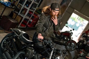 Fototapeta na wymiar Creative authentic motorcycle workshop Garage redhead bearded biker mechanic smoking cigarette near motorcycle