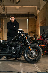 Obraz na płótnie Canvas serious bearded biker in black hoodie in real motorcycle workshop garage standing near motorcycle with crossed arms
