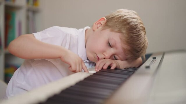 Fatigued toddler boy puts head on keyboard and presses random keys on electric piano in class closeup. Little child disliking classic music suffers during lesson
