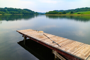 Naklejka premium Summer on Kashubia: Fishing rod and empty bucket at the calm lake