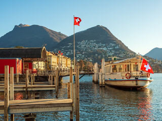 lakefront in Lugano Switzerland, with a ferry anchored at the dock, Swiss flags in the wind, and...