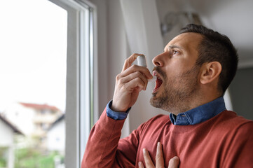 Close-up of young man in suffocation inhaling through asthma pump by window at home