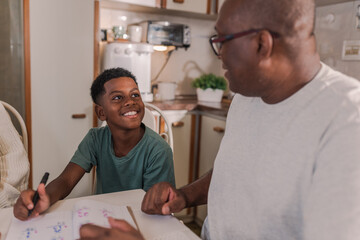 Brazilian boy sitting in the kitchen smiling to his father who is helping him with his homework in...