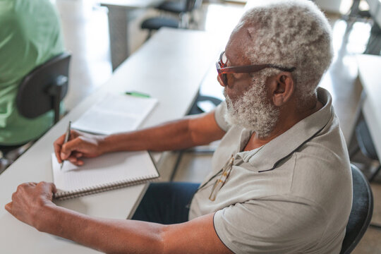 homem idoso Brasileiro negro de cabelos brancos fazendo anota&ccedil;&otilde;es no caderno durante aula em um curso para adultos no Brasil