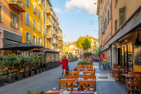 Fototapeta A young woman pushes a baby stroller down a narrow street of picturesque shops and sidewalk cafes in the colorful old town View Nice district of Nice, France, along the Cote d'Azur French Riviera.