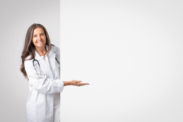 Medicine service advice. Smiling senior woman doctor in white coat pointing hand to big banner with empty space, banner