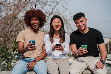 Group of multiracial teenage friends having fun using a cellphone device outdoors. Three multiethinc young people smiling and laughing watching a mobile phone sharing on social media at the street
