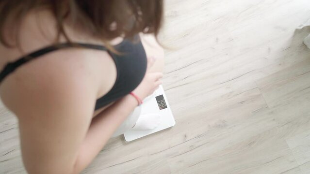 Young pregnant woman with big belly getting on floor digital scales un the home living room. Woman's health or happy pregnancy concept 4K top view footage.
