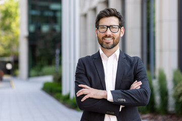 Portrait of mature adult businessman, senior boss with beard smiling and looking at camera, experienced and successful man in business suit outside office building with crossed arms - obrazy, fototapety, plakaty