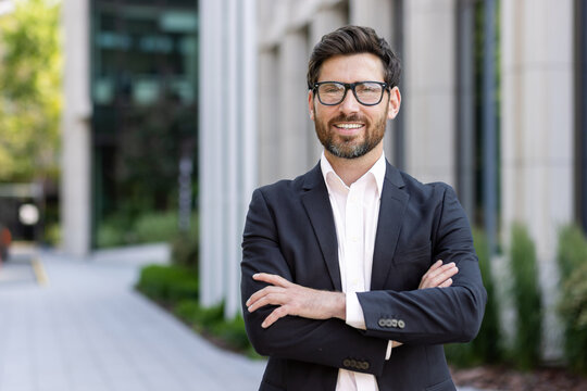 Portrait of mature adult businessman, senior boss with beard smiling and looking at camera, experienced and successful man in business suit outside office building with crossed arms