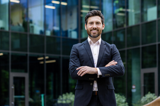 Portrait of mature adult businessman, senior boss with beard smiling and looking at camera, experienced and successful man in business suit outside office building with crossed arms