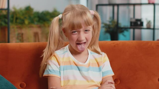 Caucasian joyful preteen school girl making playful silly facial expressions and grimacing, fooling around, showing tongue. Young excited blonde child kid at home room apartment sitting on couch
