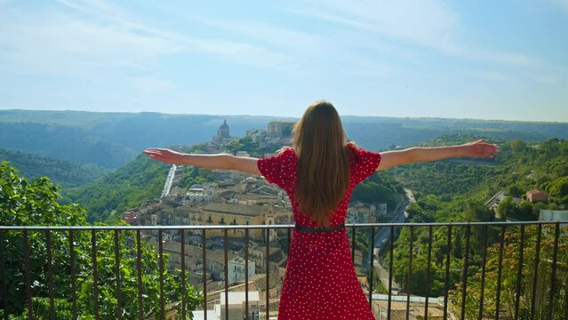 A female tourist in a beautiful red dress enjoying freedom and looking over Ibla. A young woman spreading hands towards Ragusa overlooking Ibla, Sicily.