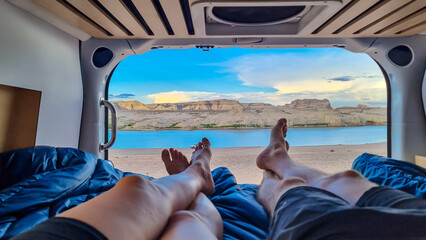 Legs of couple lying in camper van with panoramic sunset view of Wahweap Bay at Lake Powell, Glen Canyon Recreation Area, Page, Utah, USA. Sand beach on wild campground. Road trip romantic atmosphere