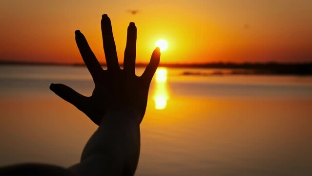 Silhouette hand of a young girl stretches out to the sunset, slow motion. Setting sun rays between fingers woman hand. Palm on the sun, faith in god. Concept dream, happiness, freedom, mental wellness