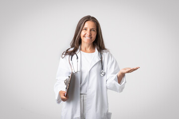 Excited senior woman doctor holding clipboard and showing something on empty palm, advertising poduct or clinic