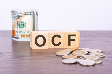top view of wooden cubes with text OCF over US dollar banknotes and coins on a brown wooden background