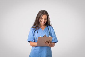 Happy senior female doc reviewing patient's medical history and taking notes to clipboard, light grey background - obrazy, fototapety, plakaty