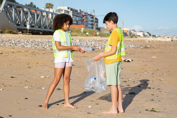 Sad serious black, arab teen pupils volunteers in vests collect garbage and bottles on beach