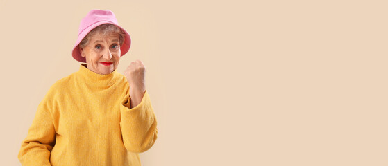 Senior woman in bucket hat showing clenched fist on beige background with space for text © Pixel-Shot