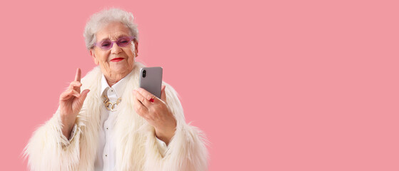 Stylish senior woman in fur coat using mobile phone on pink background with space for text