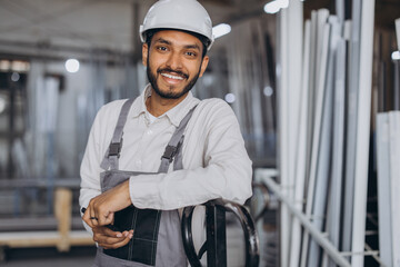 Portrait of a happy Hindu worker in a white hard hat and overalls holding a hydraulic truck against a background of a factory and aluminum frames.