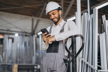 Portrait of a happy Hindu worker in a white helmet and overalls holding a hydraulic truck and talking on the phone against a background of a factory and aluminum frames.
