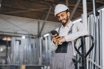 Portrait of a happy Hindu worker in a white helmet and overalls holding a hydraulic truck and talking on the phone against a background of a factory and aluminum frames.