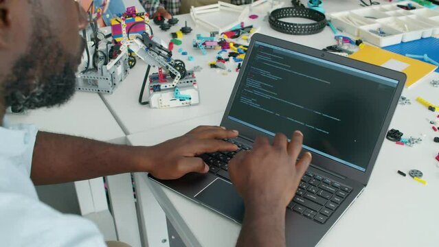 Close-up tilt down shot of African American male robotics teacher sitting at table in classroom of kids hobby club and writing computer code on laptop, and children making robot models in background