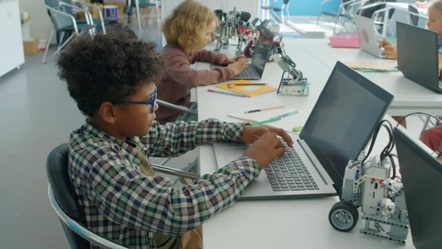 Medium side shot of preteen African American boy sitting at table with diverse classmates in after school robotics club and writing computer software for operating robot models on laptopMedium side sh