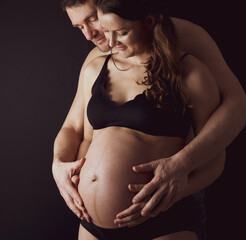 Proud parents holding naked round belly of pregnant mother in underwear. Last month of pregnancy - 36th week. Frontal view. Black background.