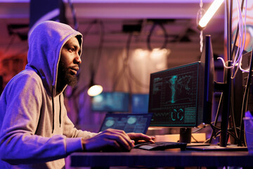 Hacker in hood breaching company cybersecurity system on computer. Young african american man breaking law while cracking password and using internet virus malicious software