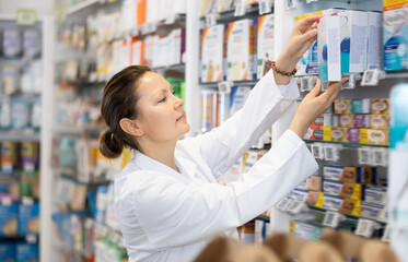 Adult woman pharmacist in uniform arranges products on shelves in pharmacy..