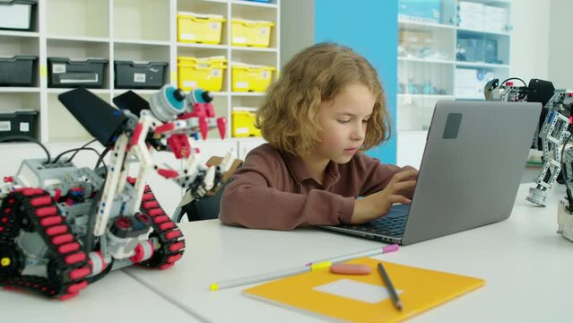 Medium shot of nine-year-old Caucasian boy sitting at table in after school IT club, looking at electric robot model and writing software to control and operate toy on laptop