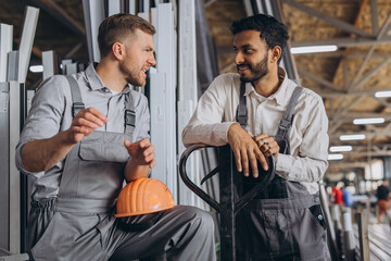 Portrait of two international workers wearing hardhats taking break from work and resting speaking to each other on a factory background with copy space
