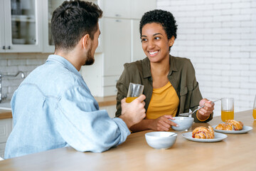 Fototapeta na wymiar Weekend morning. Happy young family couple, caucasian man and african american woman, having breakfast at cozy home kitchen with healthy porridge, happy to spend time together, communicating, smile