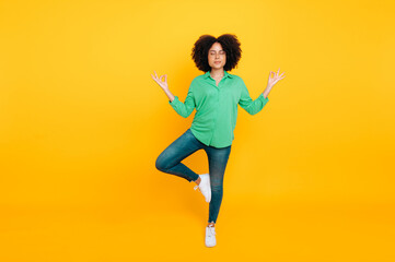 Full length photo of a stylish lovely calm smiling african american or brazilian curly woman meditating while standing tree pose with closed eyes on isolated yellow background. Mental health, harmony