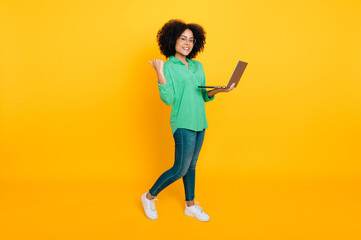 Full length photo of a stylish lovely african american or brazilian curly woman holding laptop and pointing finger back at the space for announcement, smiling, standing on isolated yellow background