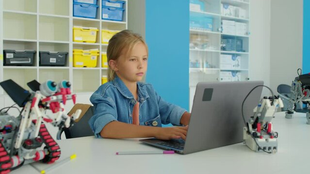 Medium shot of young Caucasian girl with blond hair, in denim shirt, sitting at table in after school kids club, looking at electric robot model, and writing computer program on laptop
