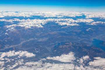 Aerial view of mountain range with snowy peaks . Clouds and blue sky as seen through window of an aircraft