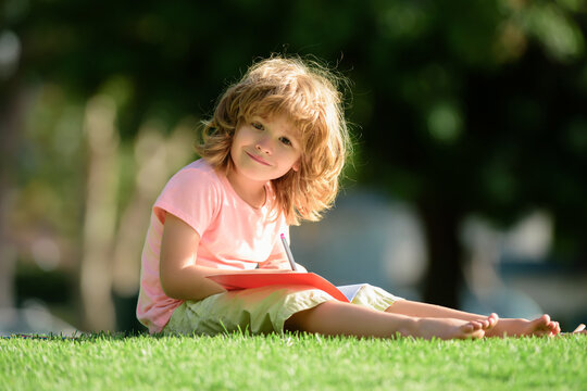 Preschool smiling pupil student boy writing to notebook in park on grass. School dream. Daydreamer school child. Dreams and imagination. Dreamy school kid face.